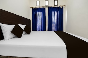 Quarto de hotel com cama, roupa de cama branca, cabeceira escura e cortinas azuis.