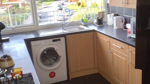 A compact kitchen with a washing machine, wooden cabinets, and a window overlooking a residential area. - Attractive apartment which has, possibly,  the best views in Felixstowe (Felixstowe)