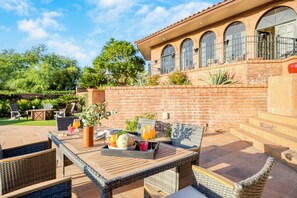 Une terrasse avec des meubles en osier, une table avec des boissons et des fruits, et un mur de briques avec une fenêtre.