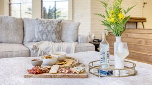 A cozy living room with a dining table set for a meal, featuring a variety of cheeses, meats, and crackers. A vase with yellow flowers and a bottle of water are also visible. - Pool, Firepit Lounge, Cornhole, and Outdoor Kitchen + Dining Area | Diamondback (Scottsdale)