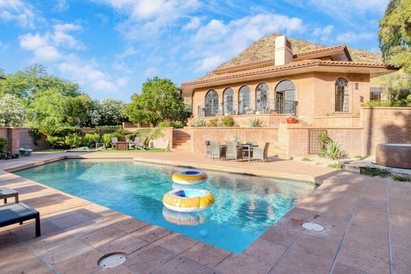 Une piscine avec deux bouées gonflables, une terrasse avec des chaises et une maison aux fenêtres en arc de cercle.