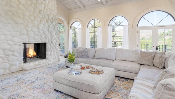 A spacious living room with a stone wall, a fireplace, a large beige sectional sofa, and arched windows. - Pool, Firepit Lounge, Cornhole, and Outdoor Kitchen + Dining Area | Diamondback (Scottsdale)