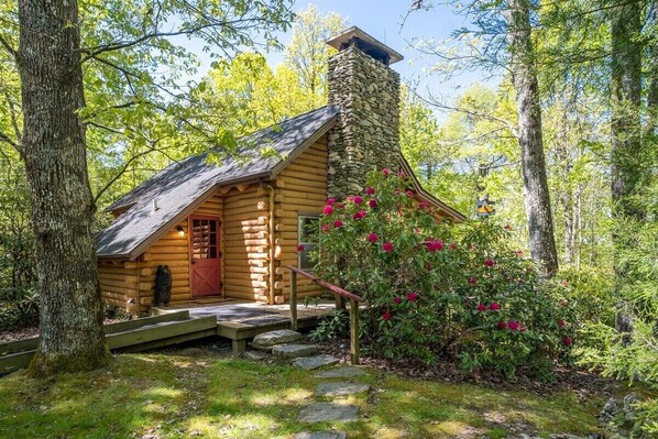 A log cabin with a stone chimney, wooden steps, and a red door.