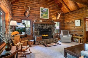 A log cabin interior with a stone fireplace, wooden walls, a mounted deer head, and a cozy seating area.