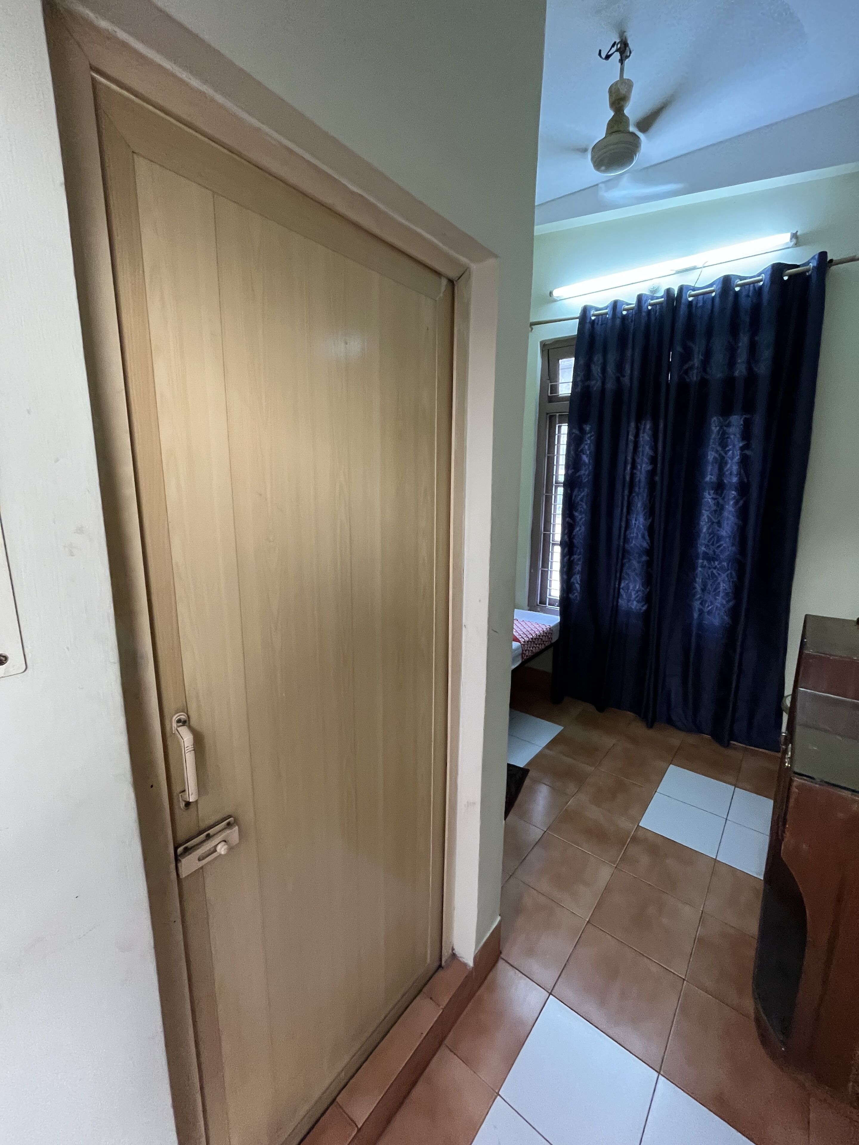 Une pièce avec une porte en bois, un lit recouvert d’une courtepointe à motifs, une fenêtre avec des rideaux et un ventilateur de plafond.