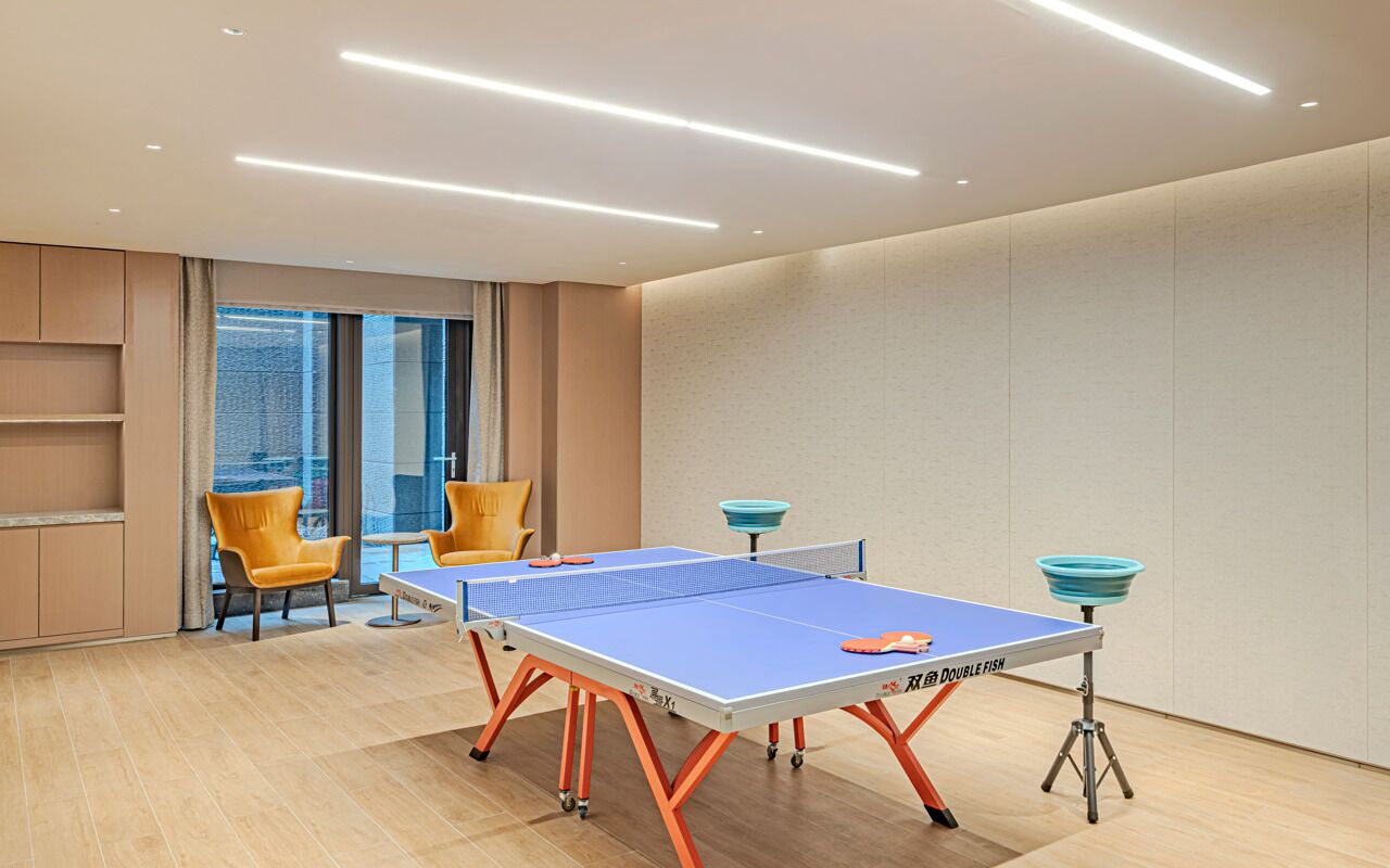 A ping pong table with a blue playing surface and orange legs, two yellow chairs, and a city view through large windows.