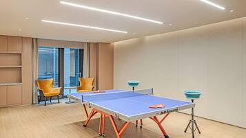 A ping pong table with a blue playing surface and orange legs, two yellow chairs, and a city view through large windows.