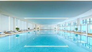 A large indoor swimming pool with clear blue water, surrounded by lounge areas with chairs and potted plants.
