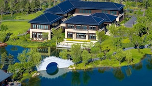 A large, modern house with a lake and a bridge, surrounded by greenery.