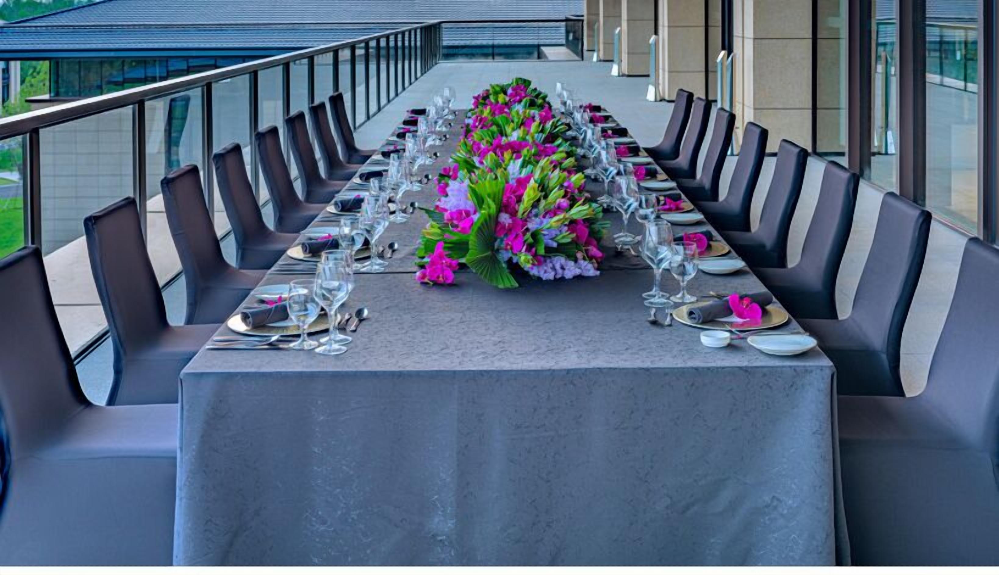 Una larga mesa de comedor dispuesta para una comida formal, con sillas a ambos lados, un centro de mesa floral y un entorno arquitectónico moderno con grandes ventanales.