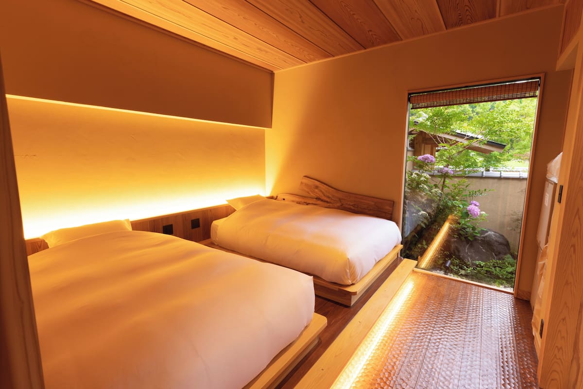 A room with two beds, wooden headboards, a small window with a view of greenery and flowers, and a wooden floor.