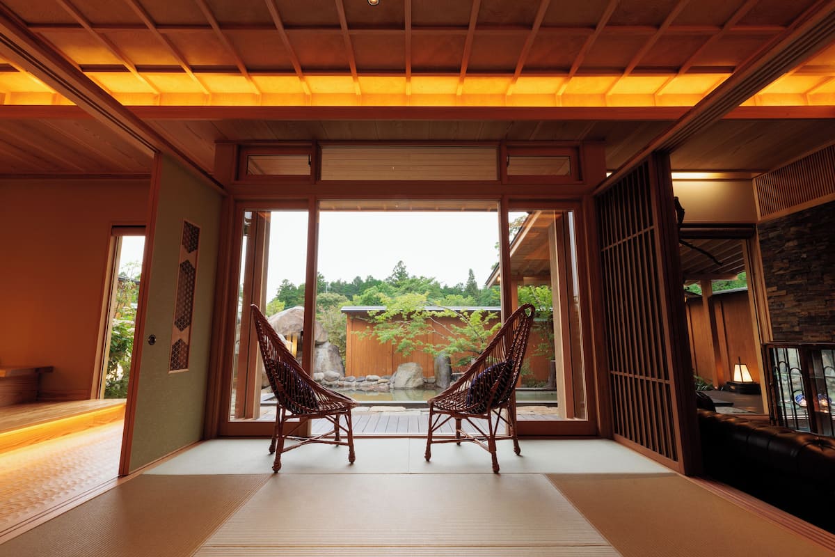 A traditional Japanese room with tatami flooring, two wooden chairs, a sliding door leading to a garden, and a view of a pond.