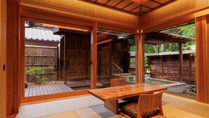 Une pièce de style japonais traditionnel, avec un sol recouvert de tatamis, une table en bois et des portes coulissantes donnant sur un espace extérieur doté d’une terrasse en bois et d’un petit étang.