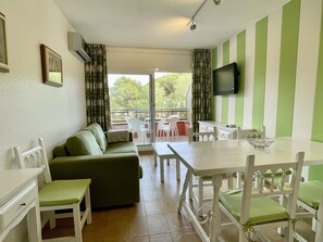 Una habitación con un sofá verde, sillas blancas, una mesa de comedor, un televisor y un balcón con sillas.