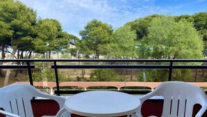 A balcony with white plastic chairs and a small round table, overlooking a beach with trees and a clear sky.