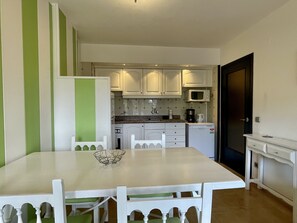 Cocina con armarios blancos, microondas y cafetera. Hay una mesa de comedor con sillas y una separador verde a rayas.