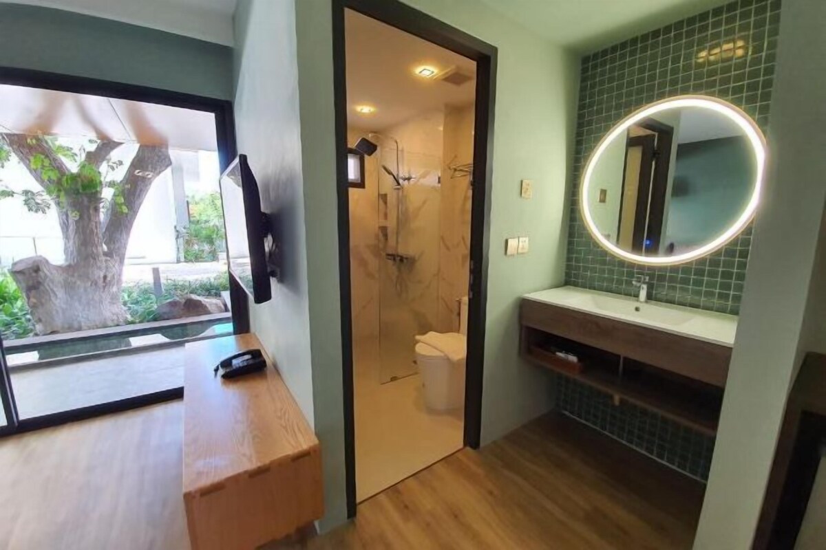 A modern bathroom with a wooden vanity, a round mirror with lights, a green-tiled wall, and a view of a tree outside.