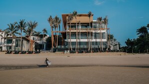 A modern building with a multi-story structure, palm trees, and a sandy beach in the foreground. - Eco Cozy Beachfront Resort Chaam (Cha-am)