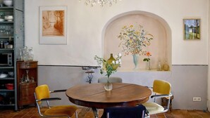 A dining area with a round wooden table, two yellow chairs, a chandelier, a painting of a tree-lined path, and a floral vase on the wall. - Authentic country house in Tannay with garden (Tannay)