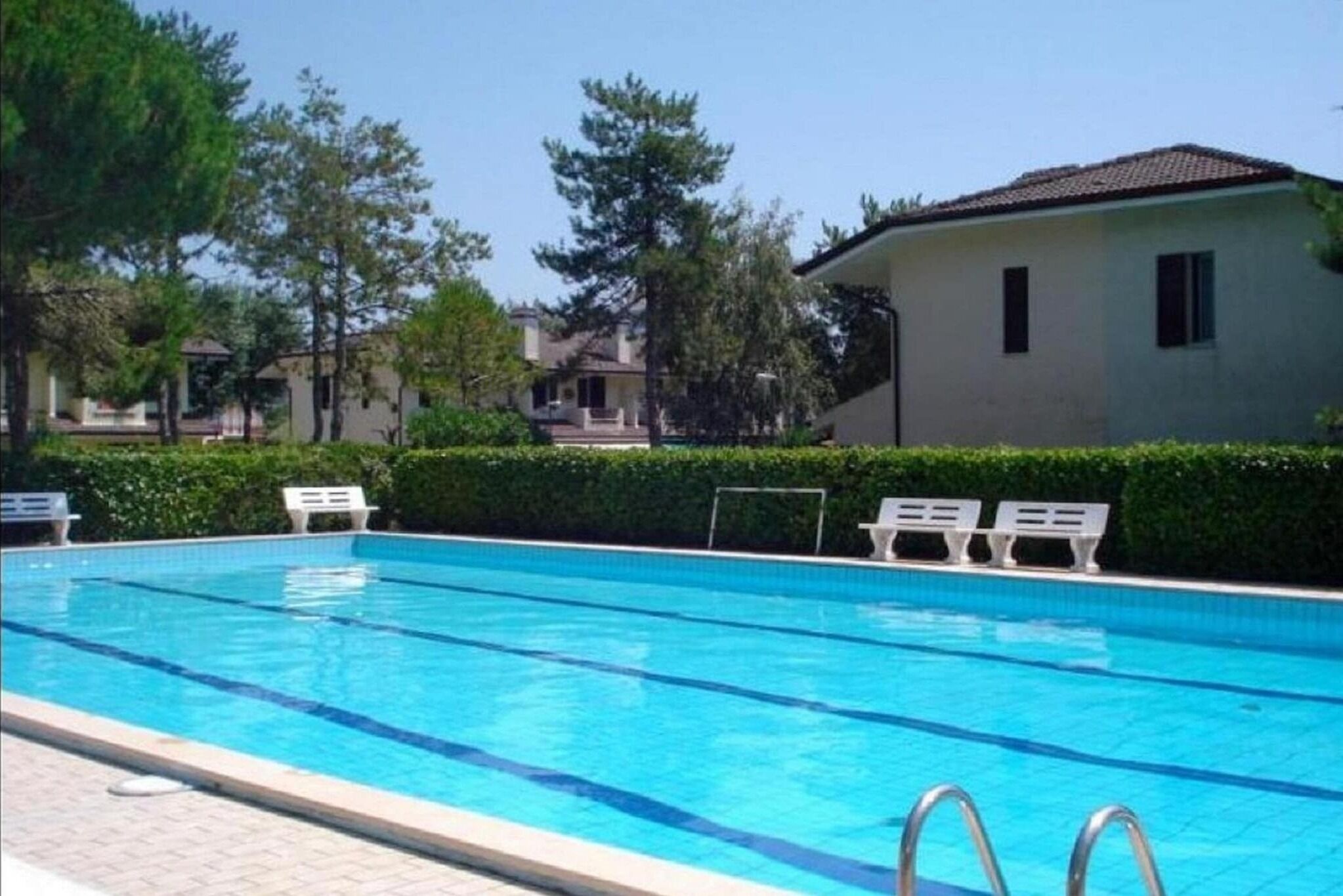 A swimming pool with clear blue water, surrounded by white lounge chairs and a hedge.