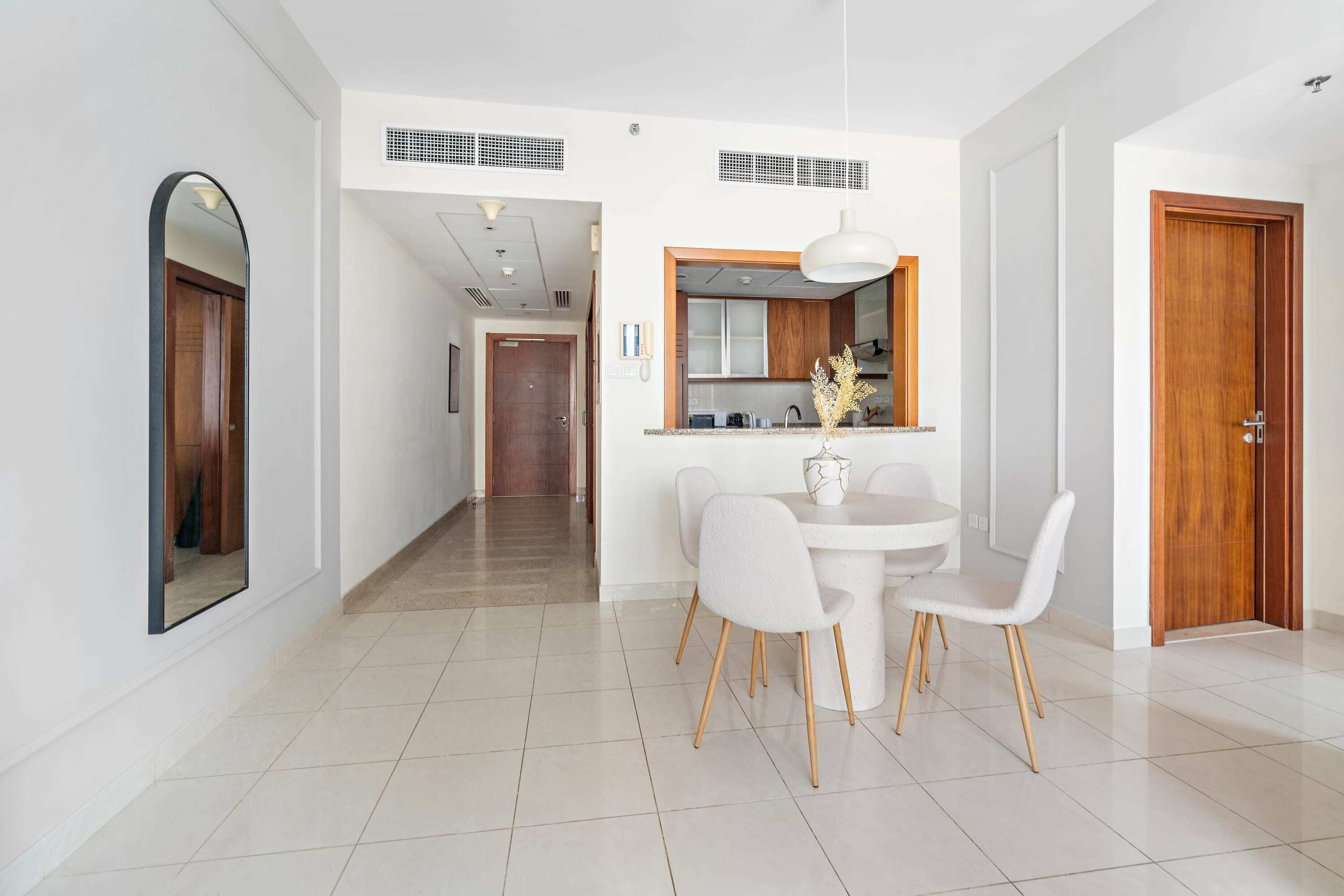 A modern dining area with a round table and white chairs, a wooden door, and a kitchen visible through an open doorway.