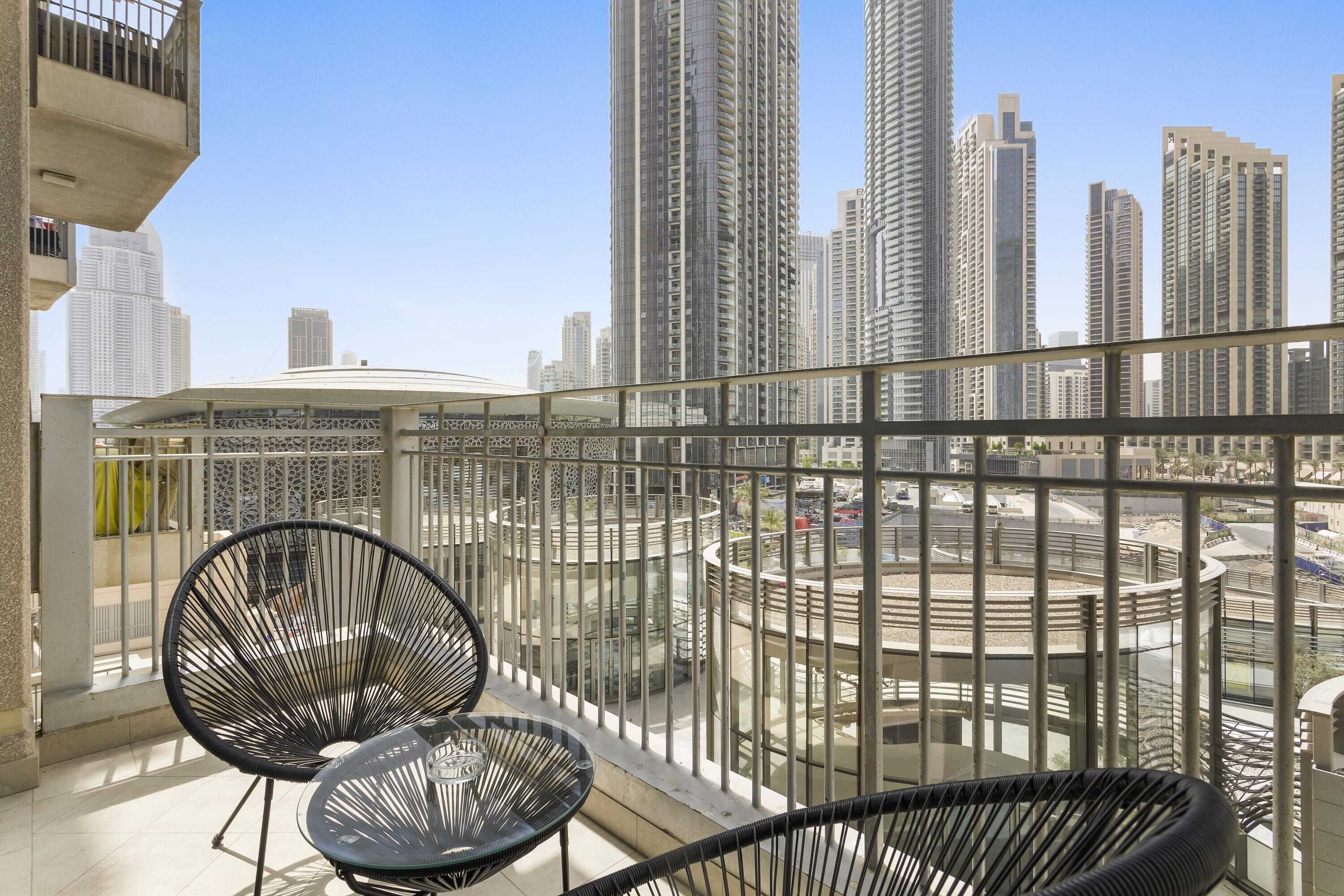 Un balcon avec deux chaises noires et une table en verre, donnant sur un paysage urbain avec de hauts immeubles et une place circulaire.