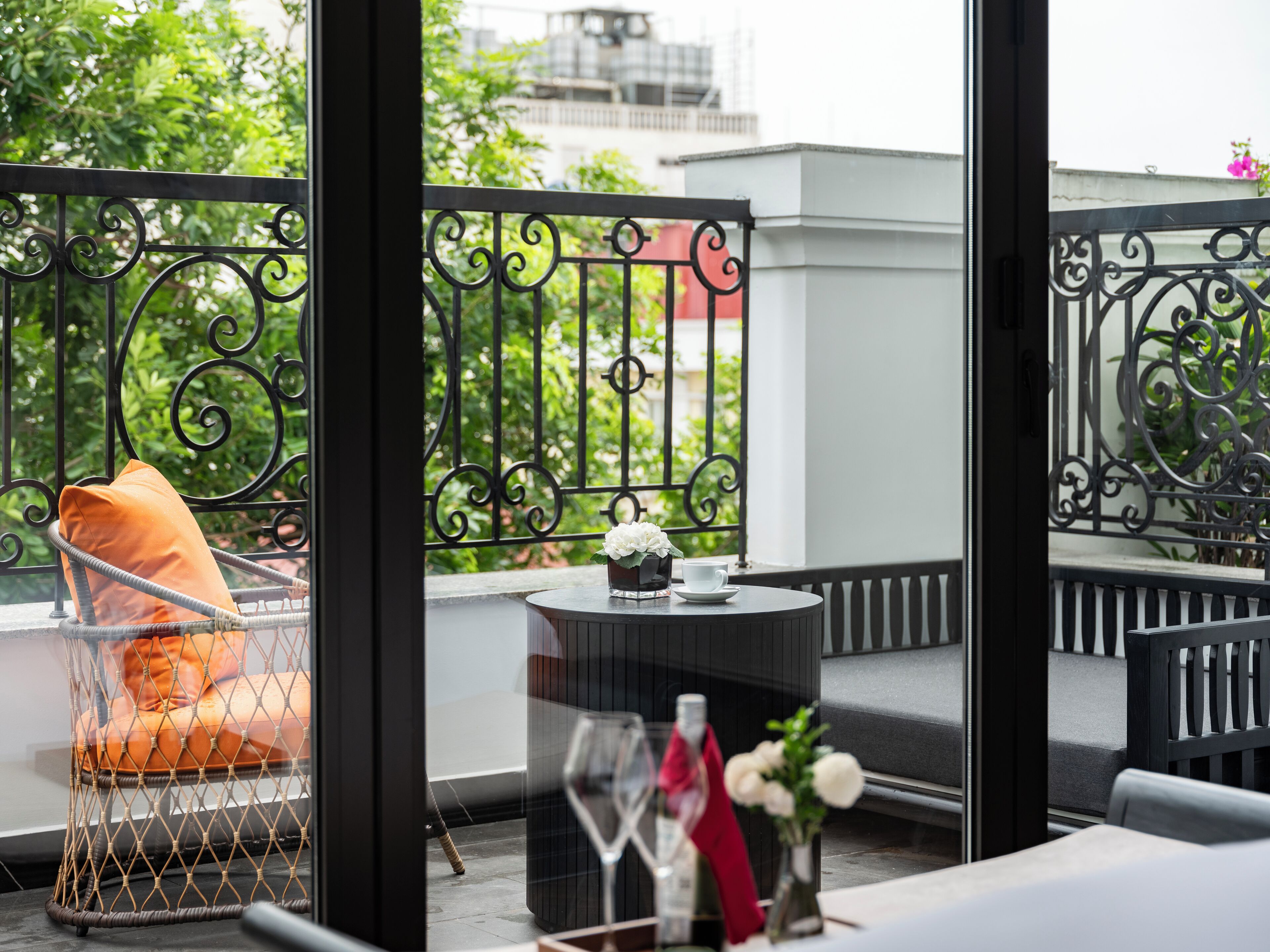 A balcony with a wicker chair, a small round table with a vase of flowers, and a glass of wine.