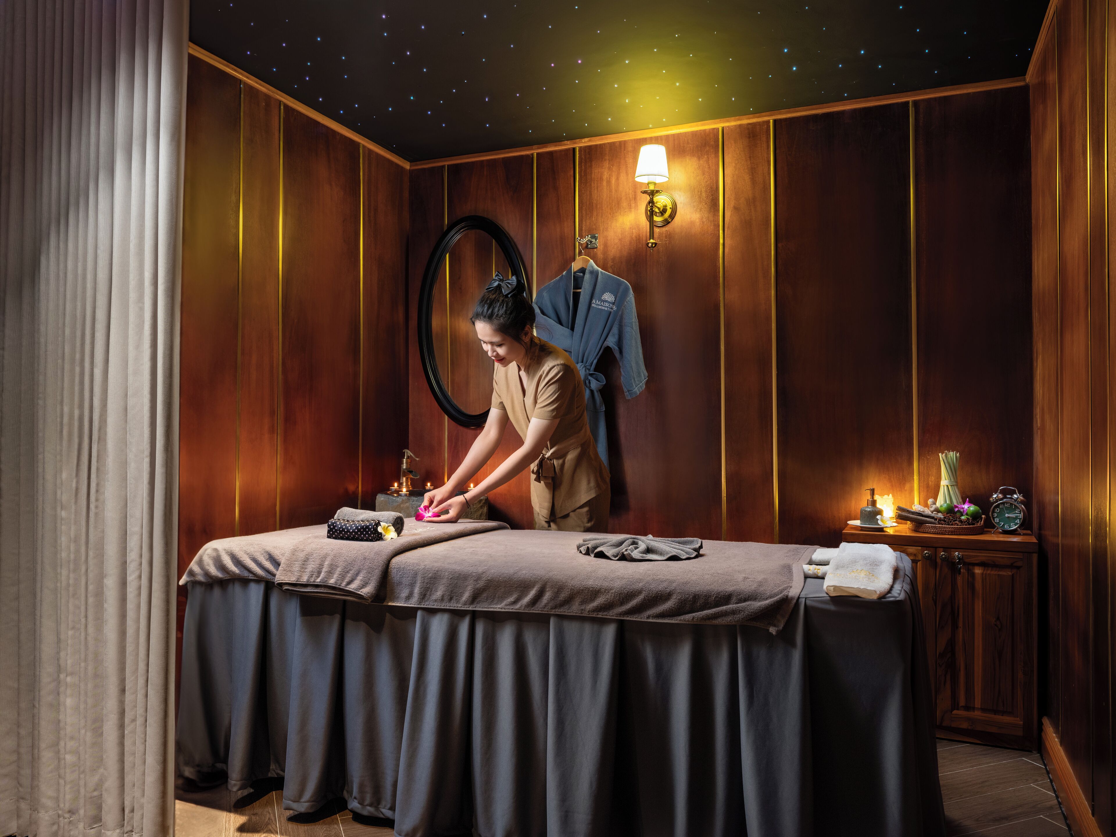 Een spa ruimte met een massagetafel, een badjas, een handdoek, een klok en een klein tafeltje met kaarsen en een fruitmand.