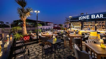 An outdoor bar area with tables and chairs, a palm tree, and a building in the background.