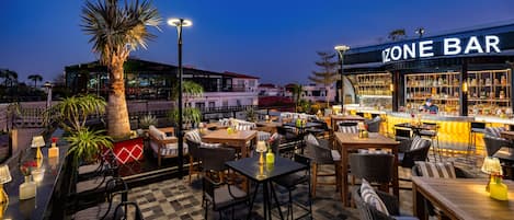 An outdoor bar area with tables and chairs, a palm tree, and a building in the background.