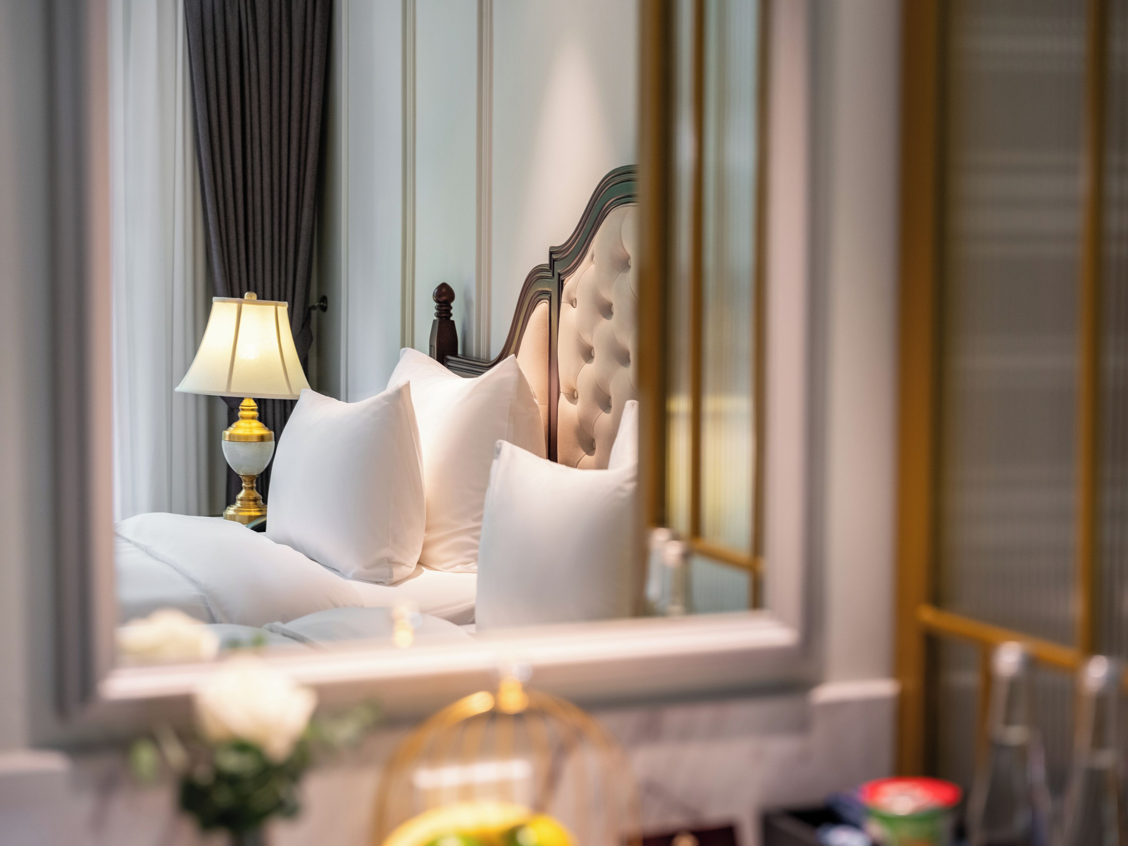 Un lit avec du linge de lit blanc et une tête de lit capitonnée, une lampe de chevet et un miroir qui reflète l’intérieur de la pièce.