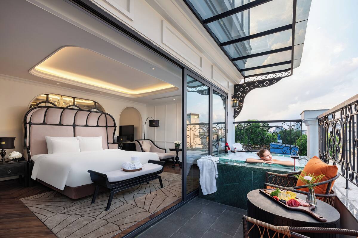 Une chambre d’hôtel luxueuse dotée d’un grand lit, d’un coin salon avec vue, d’un balcon avec jacuzzi et d’une table à manger accompagnée d’une planche de fromages.