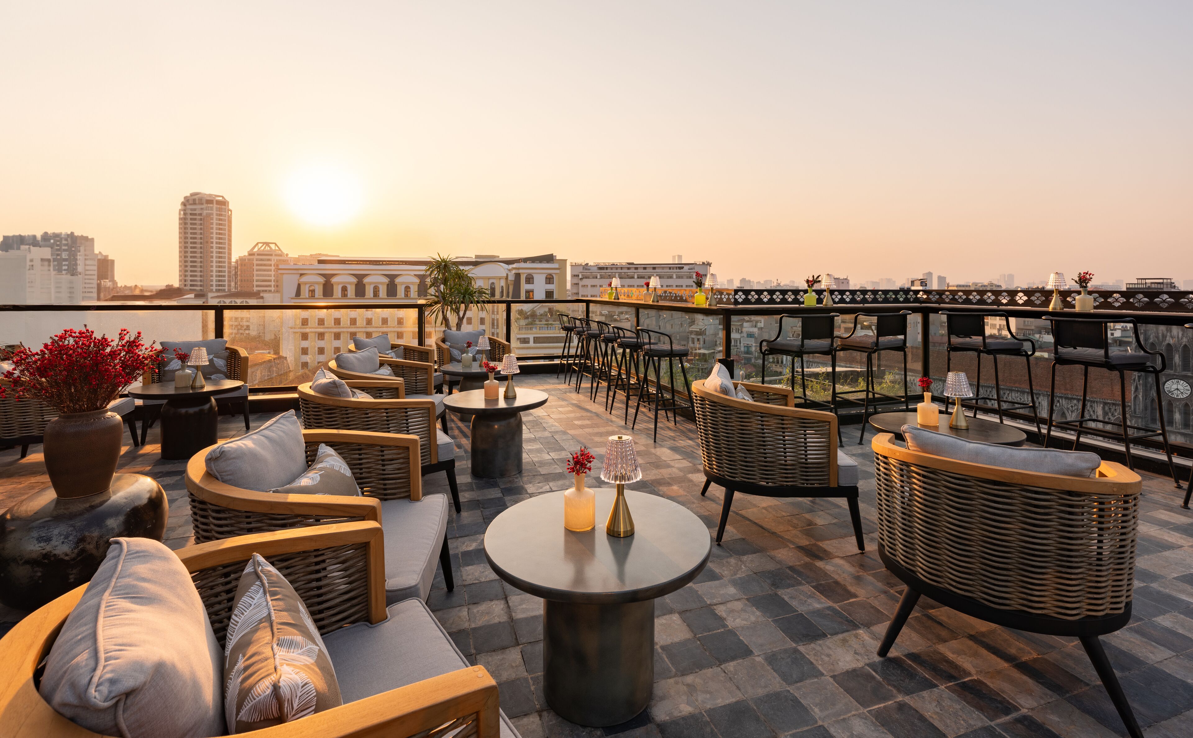 A rooftop terrace with wicker furniture, round tables, and a view of the city skyline at sunset.