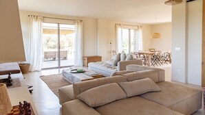 Ein großzügiges Wohnzimmer mit einem breiten, beigefarbenen Sofa, einem Holztisch und einem Schachbrett. Zwei Fenster mit weißen Vorhängen, eine Essecke mit Tisch und Stühlen sowie eine Pendelleuchte.