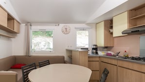 A compact kitchen with a round table, built-in cabinets, a stove, and a window with curtains. - Caravan 'Midi - Mobil Home 6-8 Pers' with Shared Terrace and Shared Garden (Vallon-Pont-d'Arc)