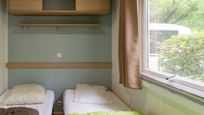A room with two beds, a wooden shelf above, and a window with a view of greenery. - Caravan 'Midi - Mobil Home 6-8 Pers' with Shared Terrace and Shared Garden (Vallon-Pont-d'Arc)