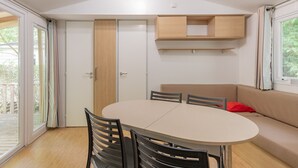 A room with a round table, chairs, a sofa, a sliding door, and built-in shelves. - Caravan 'Midi - Mobil Home 6-8 Pers' with Shared Terrace and Shared Garden (Vallon-Pont-d'Arc)