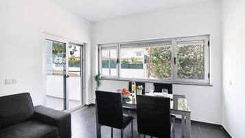 A modern room with a dining table, two chairs, a sofa, and large windows offering a view of the outdoors.