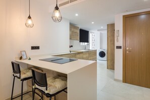 Una cocina moderna con una isla central, taburetes de bar, iluminación colgante y una lavadora al fondo.