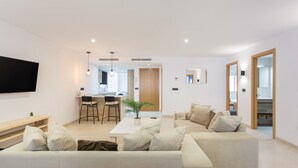 Een moderne woonkamer met een bank, een salontafel en een televisie. In de achtergrond is een keuken met een eettafel en stoelen.