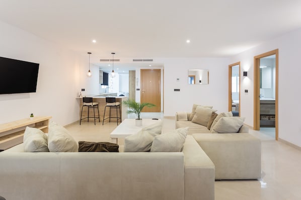 A modern living room with a sofa, a coffee table, and a TV. There is a kitchen area in the background with a dining table and chairs.