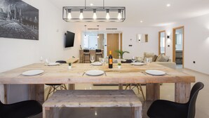 Een eetgedeelte met een houten tafel, stoelen en een bank. Er hangt een schilderij aan de muur, er is een televisie en een plafondlamp.