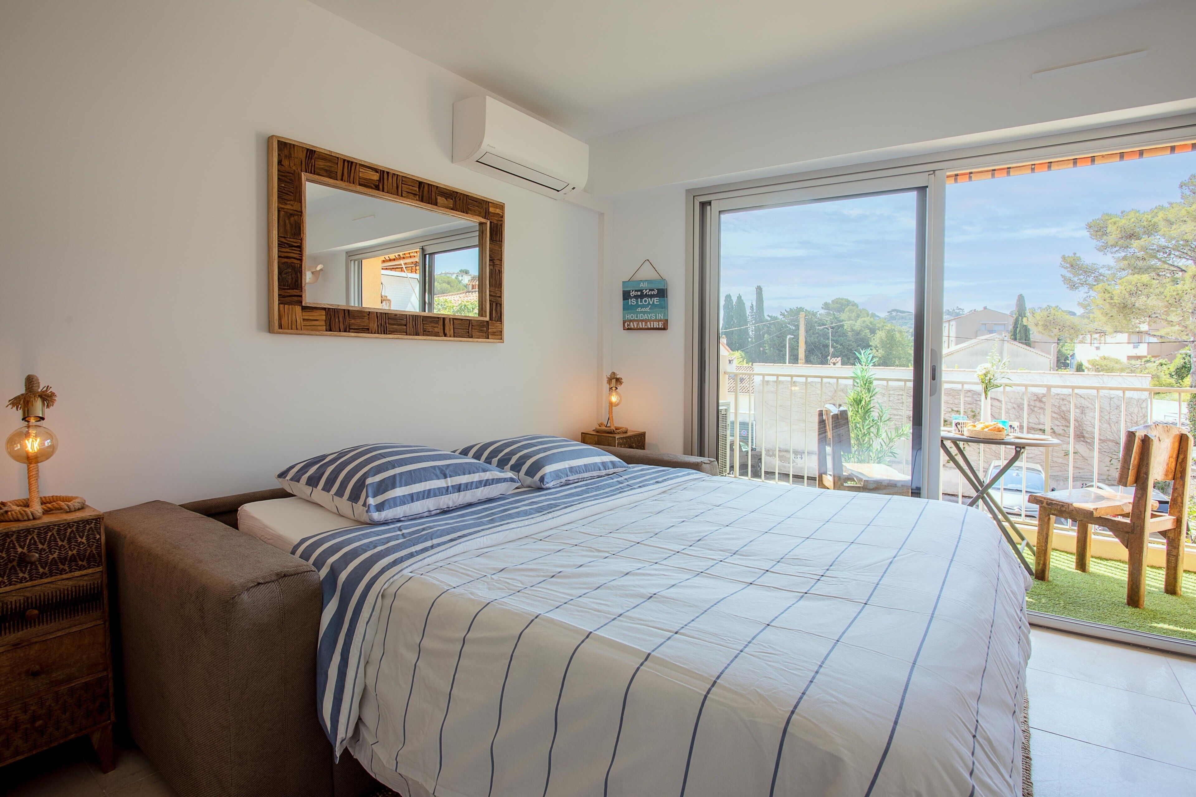 A bedroom with a bed, a nightstand, a lamp, a mirror, and a view of the outdoors.