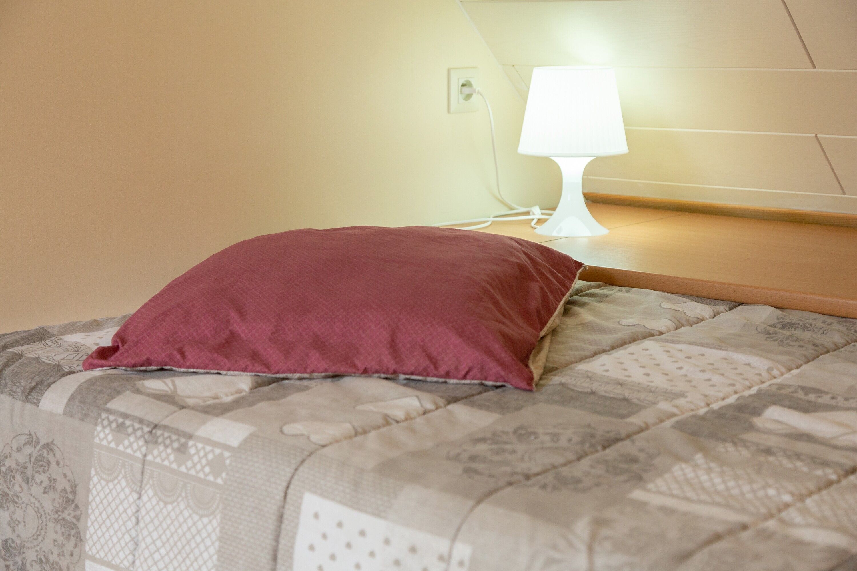 A bed with a patterned bedspread and a single red pillow.