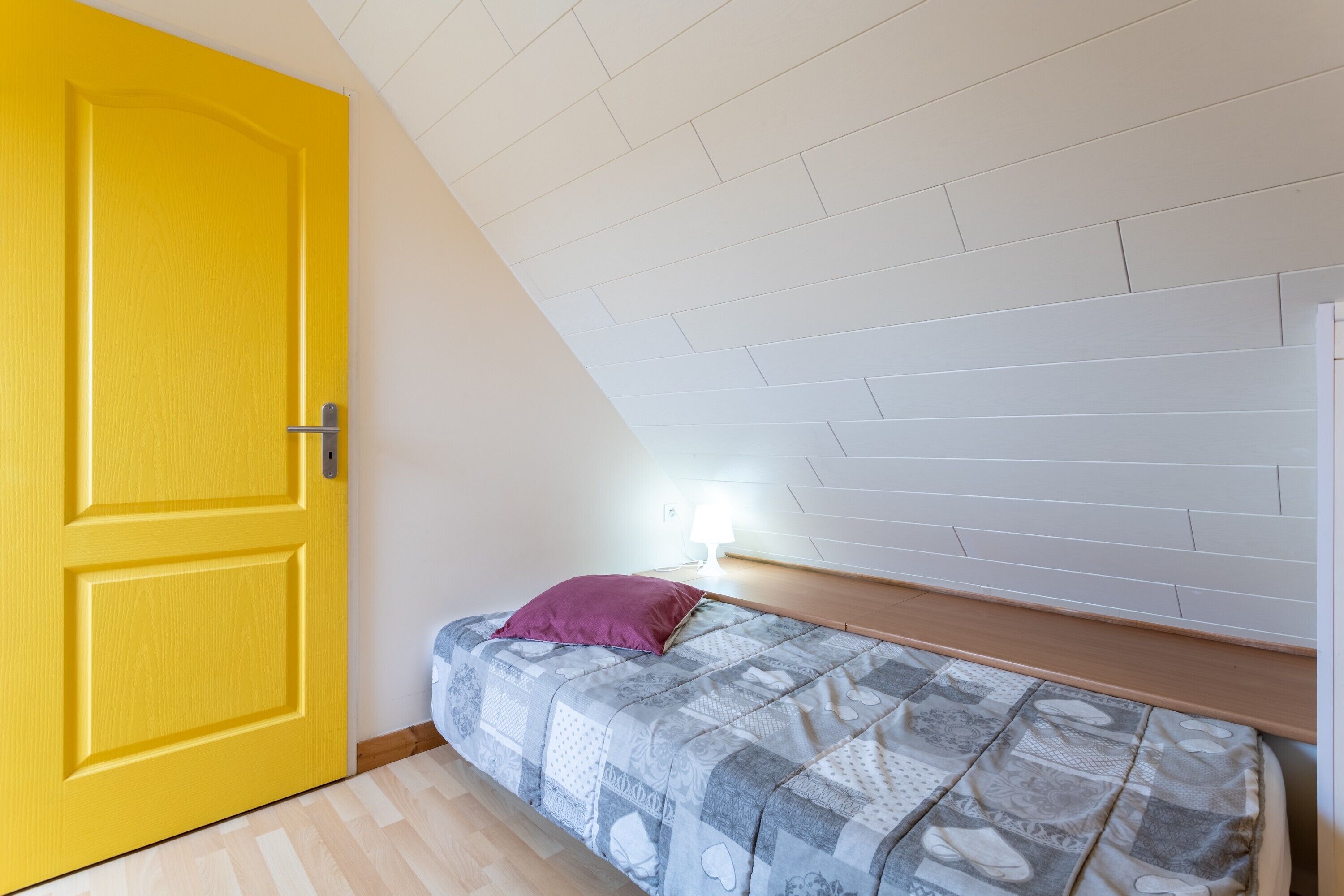 Un dormitorio con techo inclinado, una cama con colcha estampada, una puerta amarilla y una mesita de noche con lámpara.