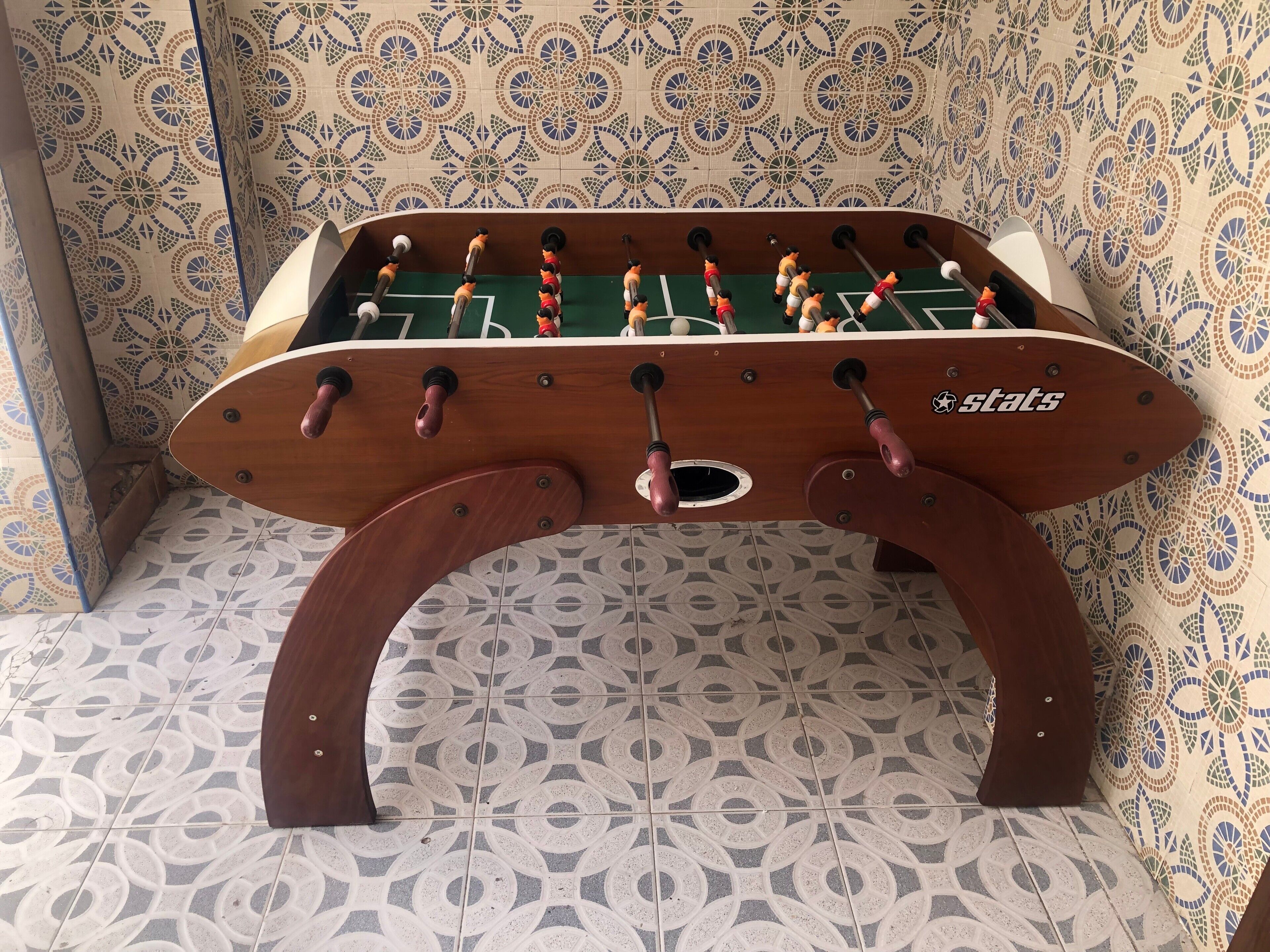 Ein Kicker mit Holzfuß und Rückenlehne vor einer Wand mit Muster.