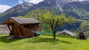Una cabaña de madera con techo inclinado, una hamaca y un muro de piedra en una zona montañosa.