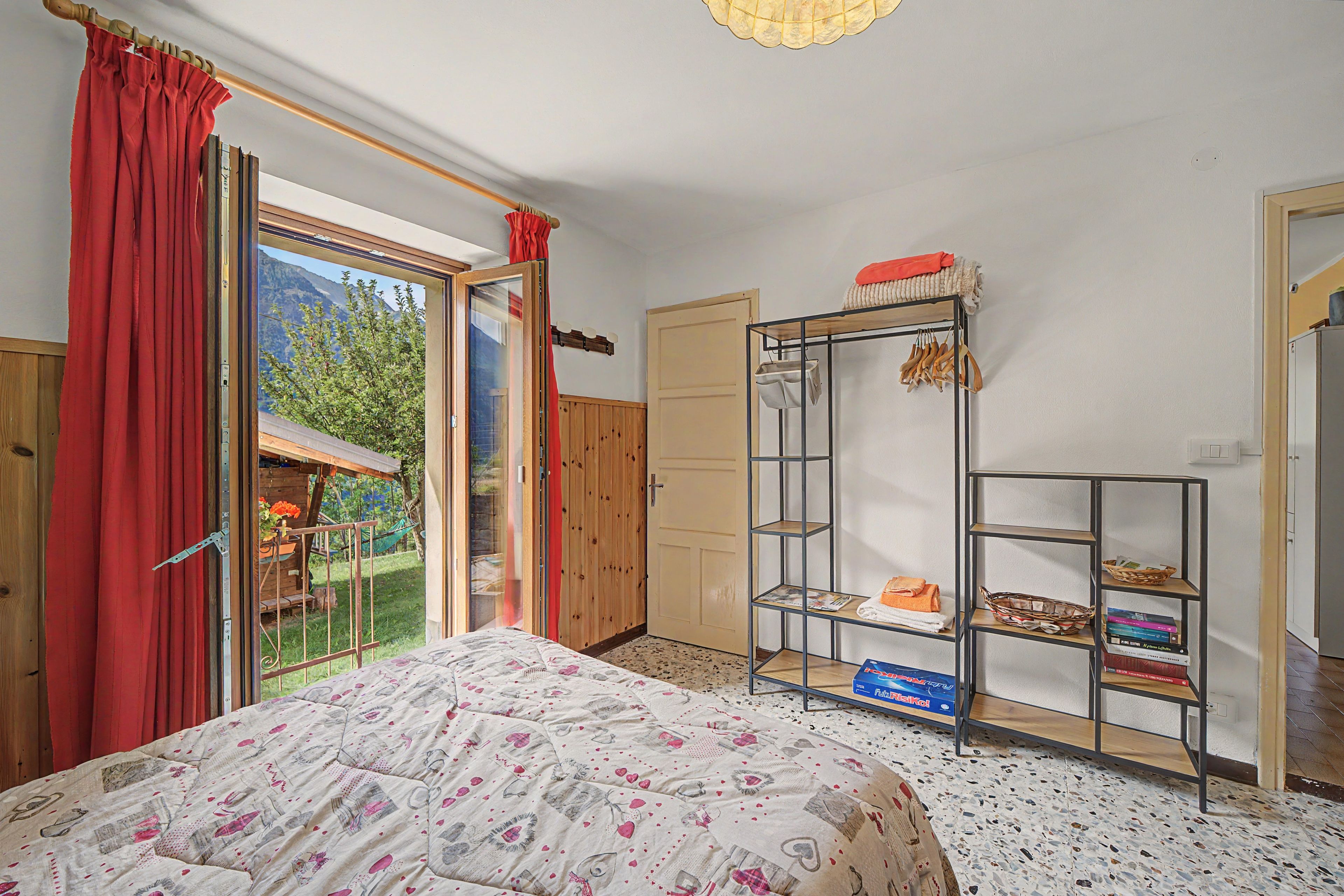Ein Schlafzimmer mit einem Bett, einem Kleiderschrank, einer Ablage mit Handtüchern und Büchern sowie einem Blick auf einen Garten durch ein Fenster.