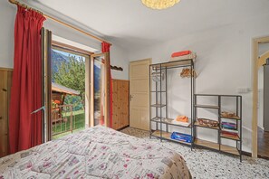 Une chambre à coucher comprenant un lit, une armoire, une étagère avec des serviettes et des livres, et donnant sur un jardin par une fenêtre.