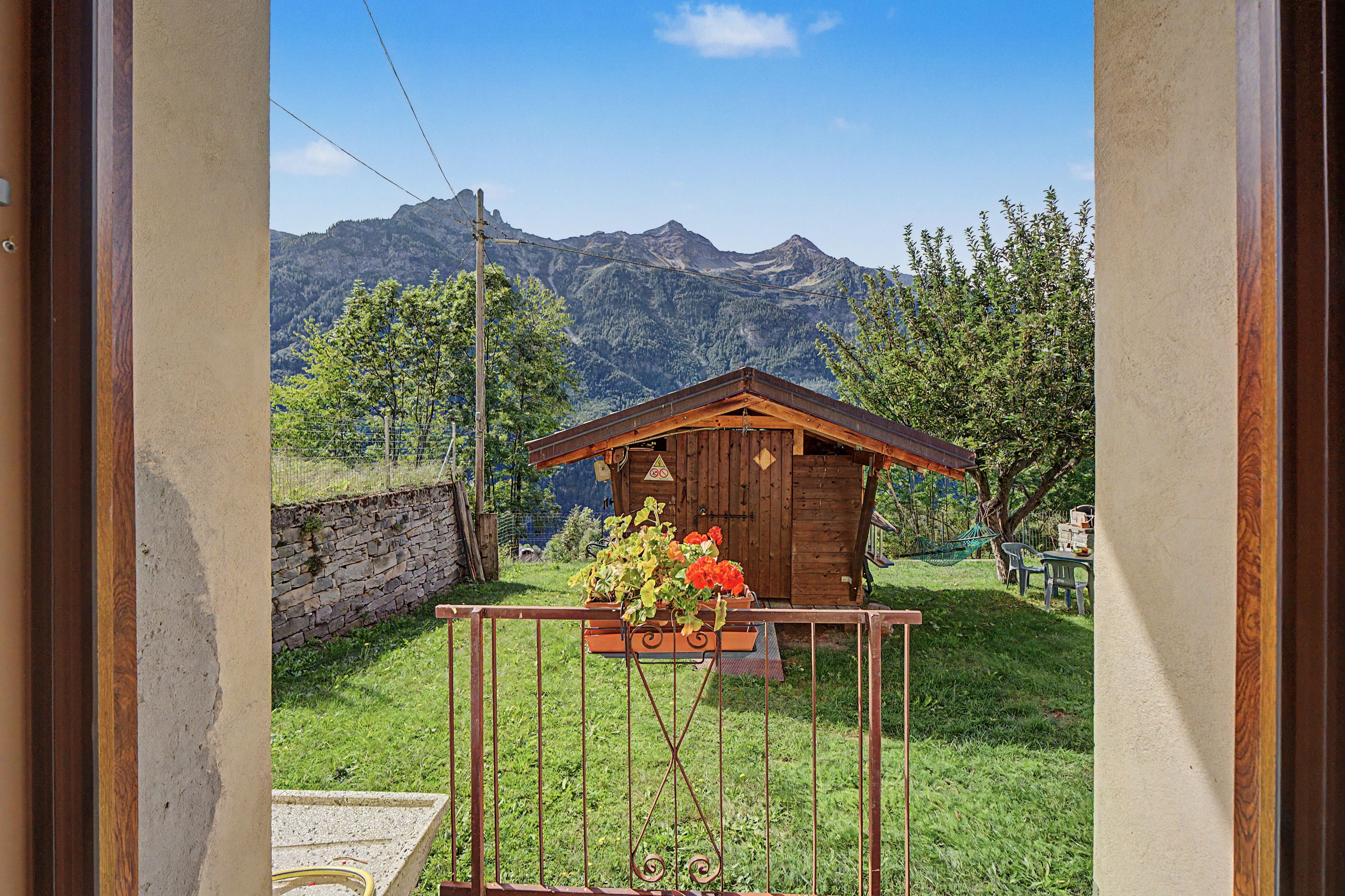 Una baita di legno con parete in pietra e cancello in metallo, immersa nel verde con montagne sullo sfondo.